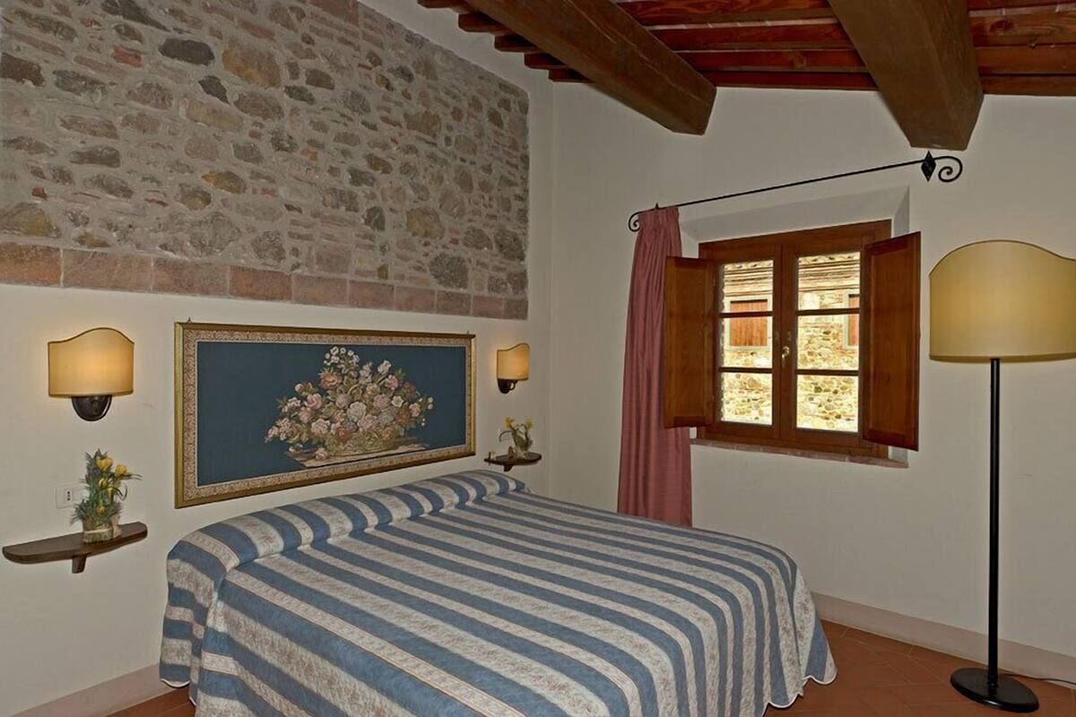 Une chambre avec un mur en pierre, un lit recouvert d’une couvre-lit à rayures, un tableau floral encadré, deux lampes fixées au mur, une fenêtre avec un rideau et une lampe sur pied.