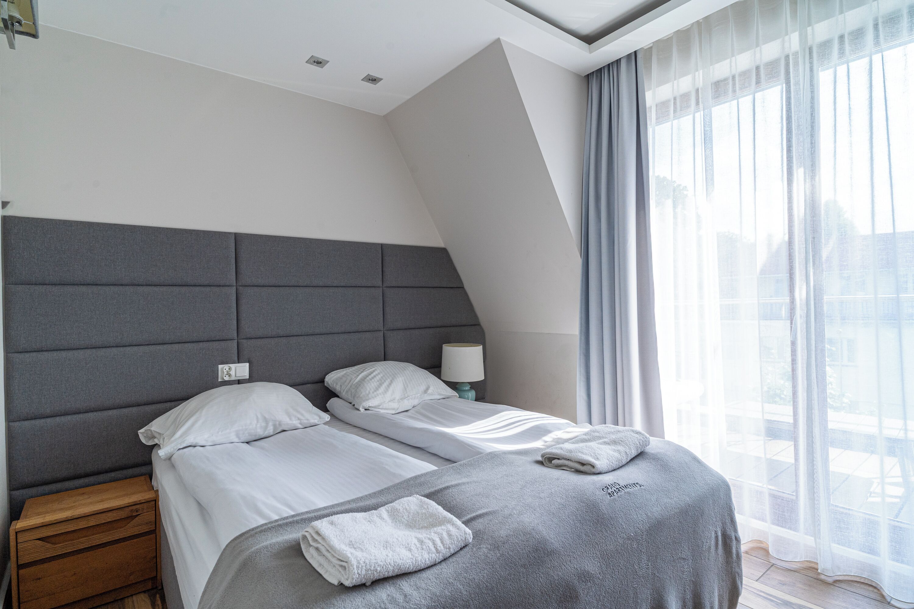 Ein modernes Hotelzimmer mit einem großen Bett, einem Nachttisch und einem Fenster mit durchsichtigen Vorhängen.