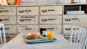 A table with a croissant, a glass of orange juice, and a cup of coffee. Behind the table, there are white chairs and a cabinet with drawers.