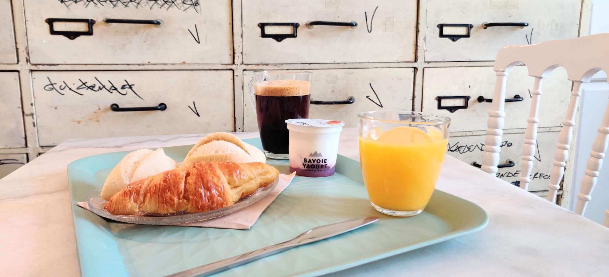 A breakfast tray with croissants, a glass of orange juice, and a cup of yogurt on a table with a vintage-style cabinet in the background.