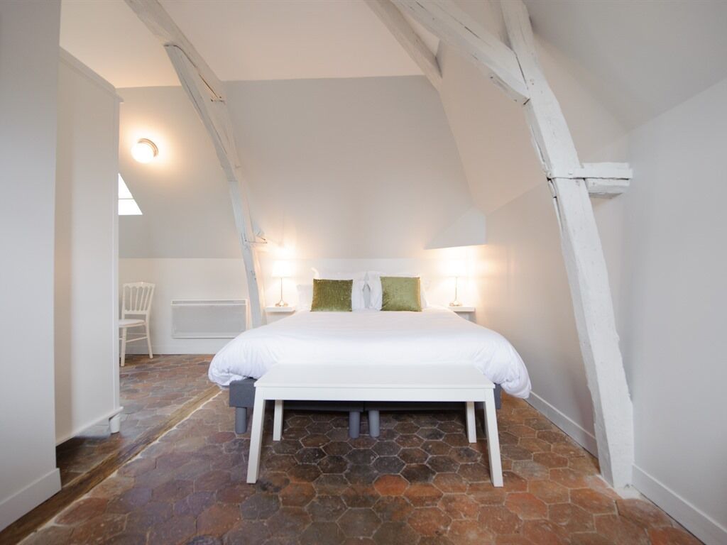 Un dormitorio con cama blanca, suelo de piedra, paredes blancas y vigas de madera a la vista.