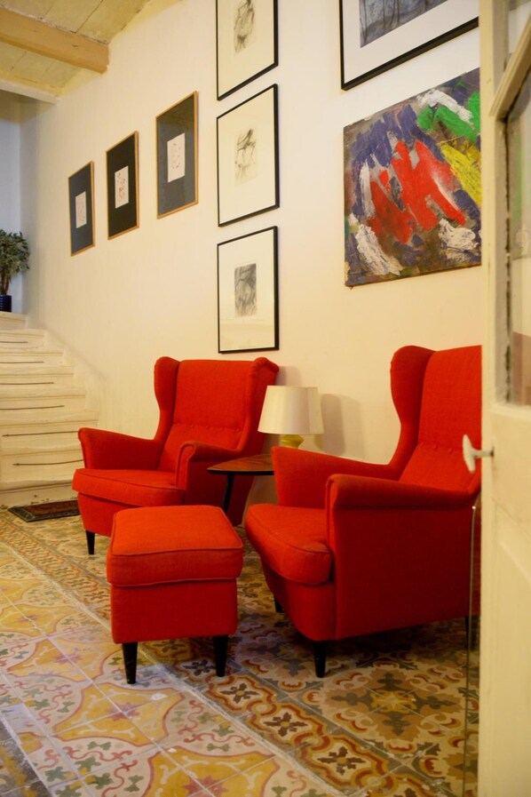 A room with a staircase, red armchairs, a small table with a lamp, and framed artwork on the wall.