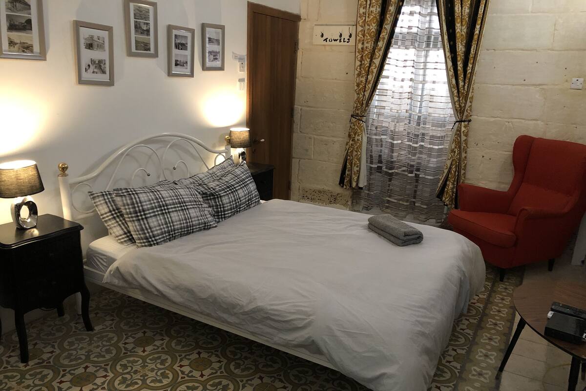 A neatly made bed with a white comforter and plaid pillows, a red armchair, a small table with a lamp, and patterned floor tiles.