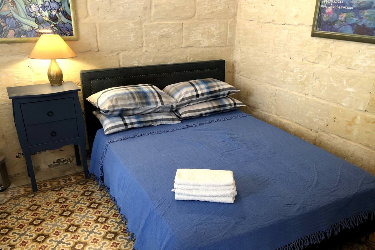 A neatly made bed with a blue bedspread and a folded white towel, a dark blue nightstand with a lamp, and a patterned floor.