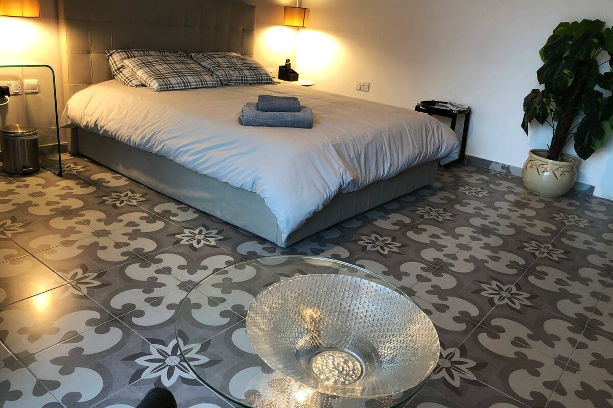 A neatly arranged bedroom with a bed, bedside lamps, a glass table, and a potted plant.