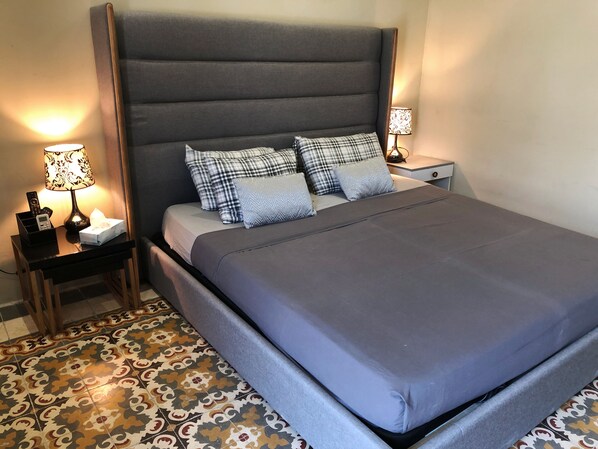 A neatly made bed with a grey headboard and multiple pillows, a bedside table with a lamp, and a patterned floor.