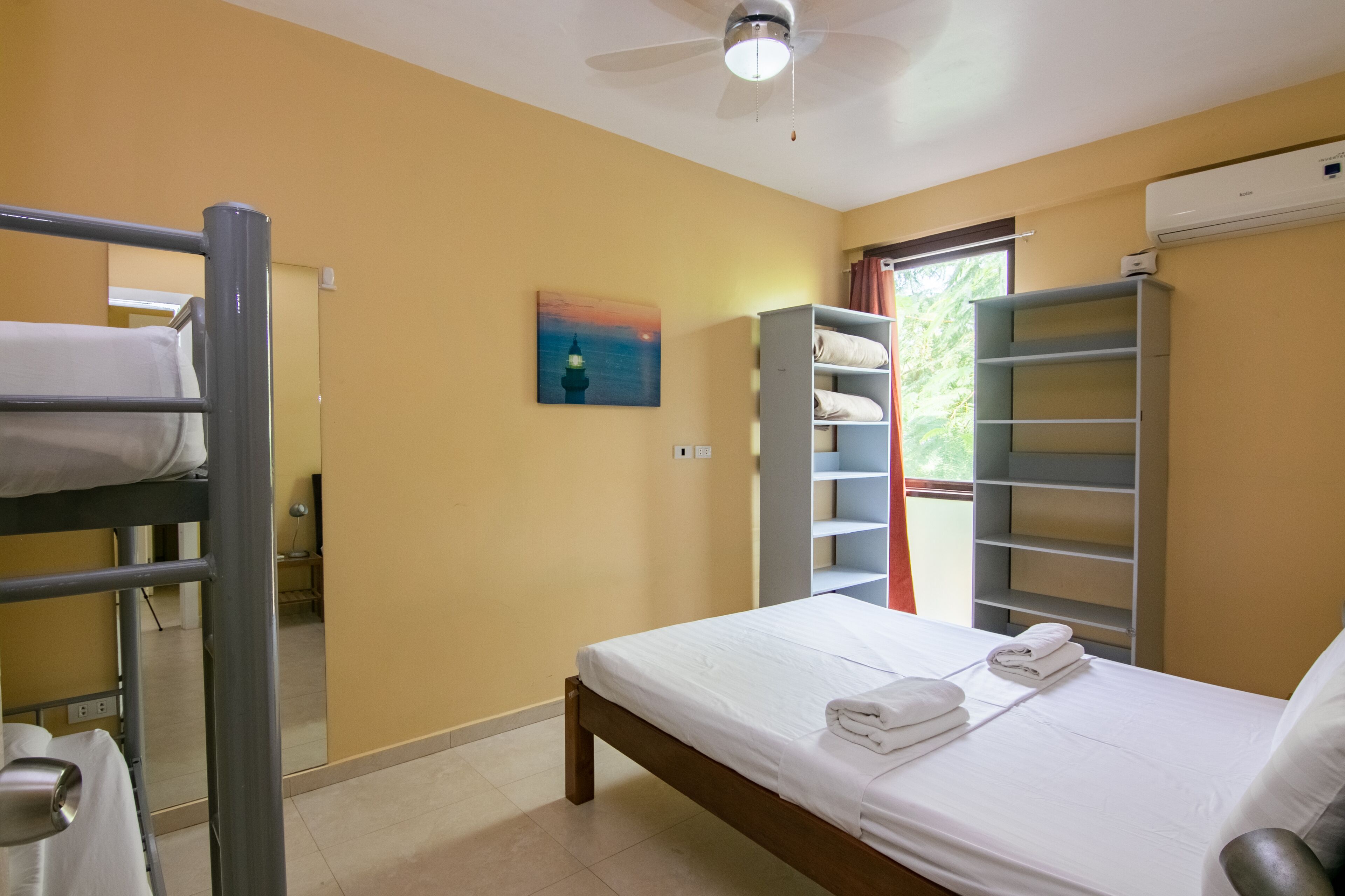 A hotel room with a bunk bed, a single bed, a painting, a wardrobe, and a window with curtains.
