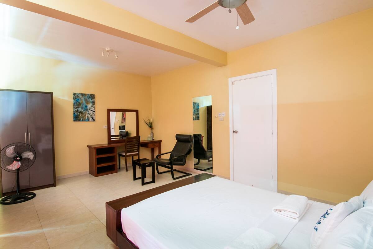 Habitación de hotel con cama, escritorio, silla, armario y ventilador de techo.