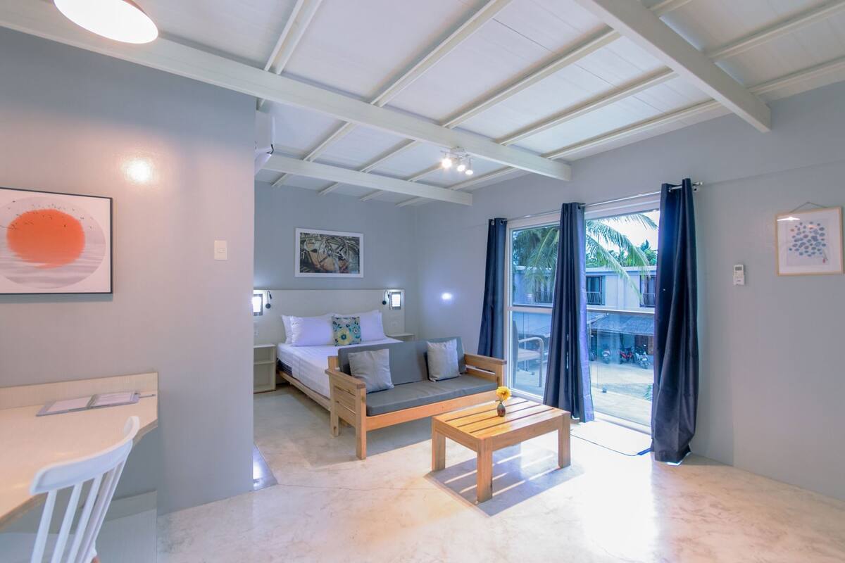 Una habitación con cama, escritorio, silla, mesita y vista por una puerta de cristal.