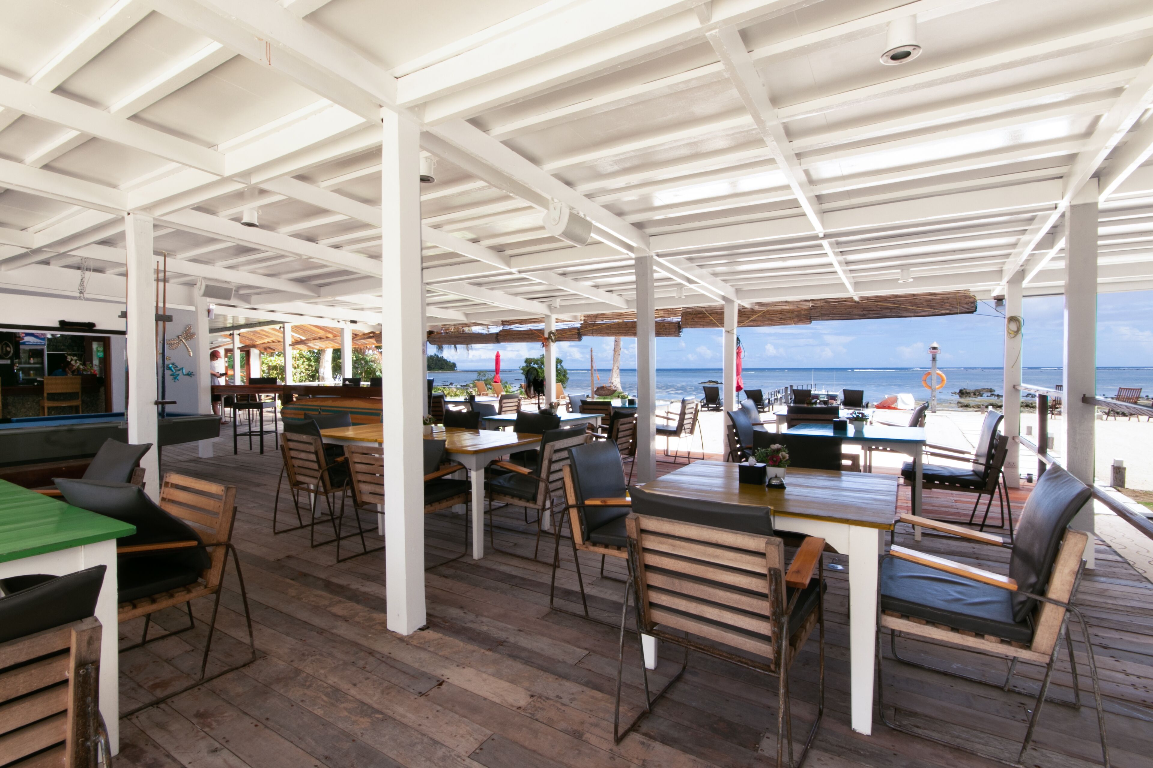 An outdoor dining area with wooden tables and chairs, a view of the ocean, and a covered structure with a white ceiling.