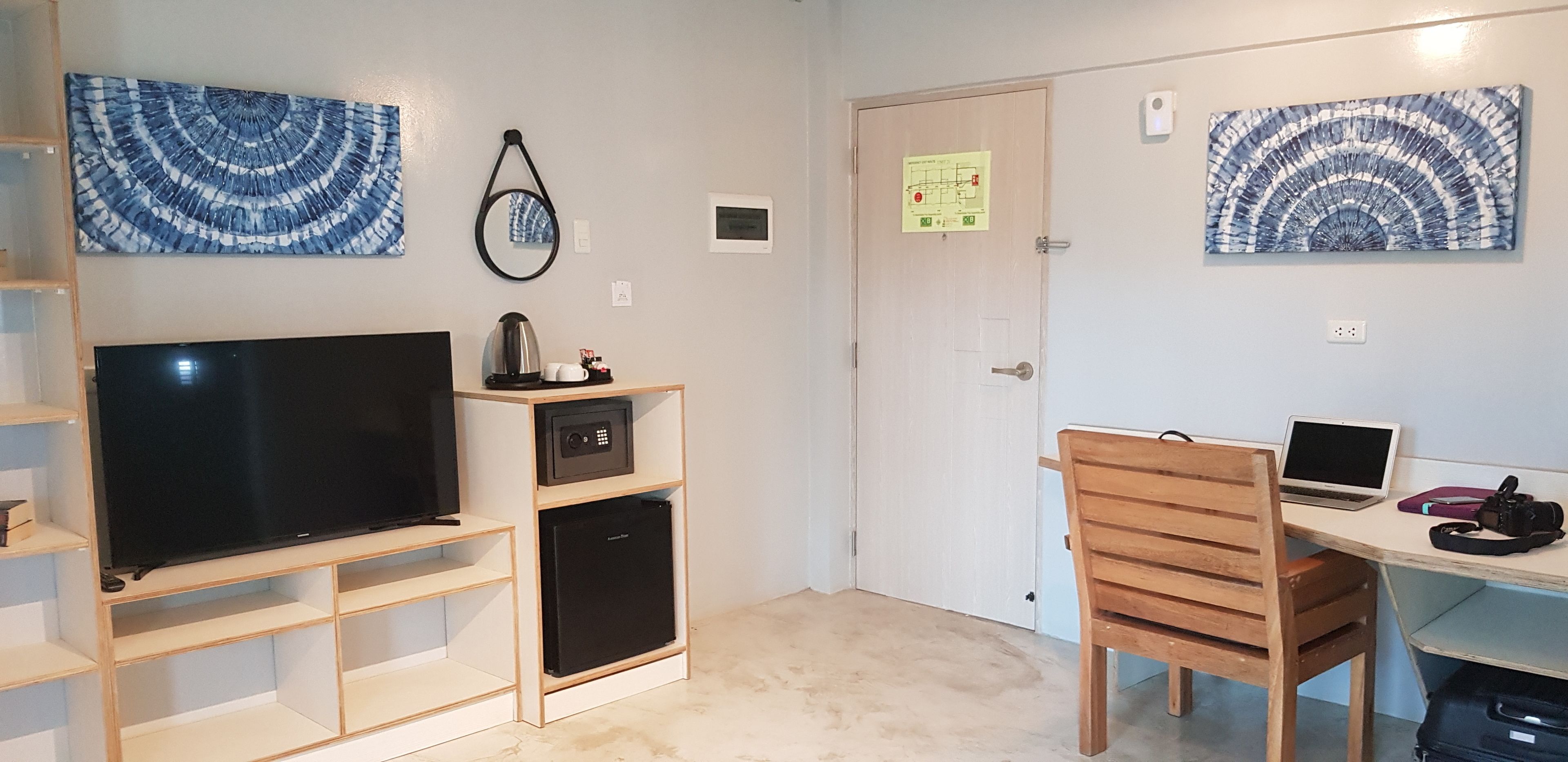 A room with a TV, a coffee machine, a refrigerator, a desk with a laptop, and two paintings on the wall.
