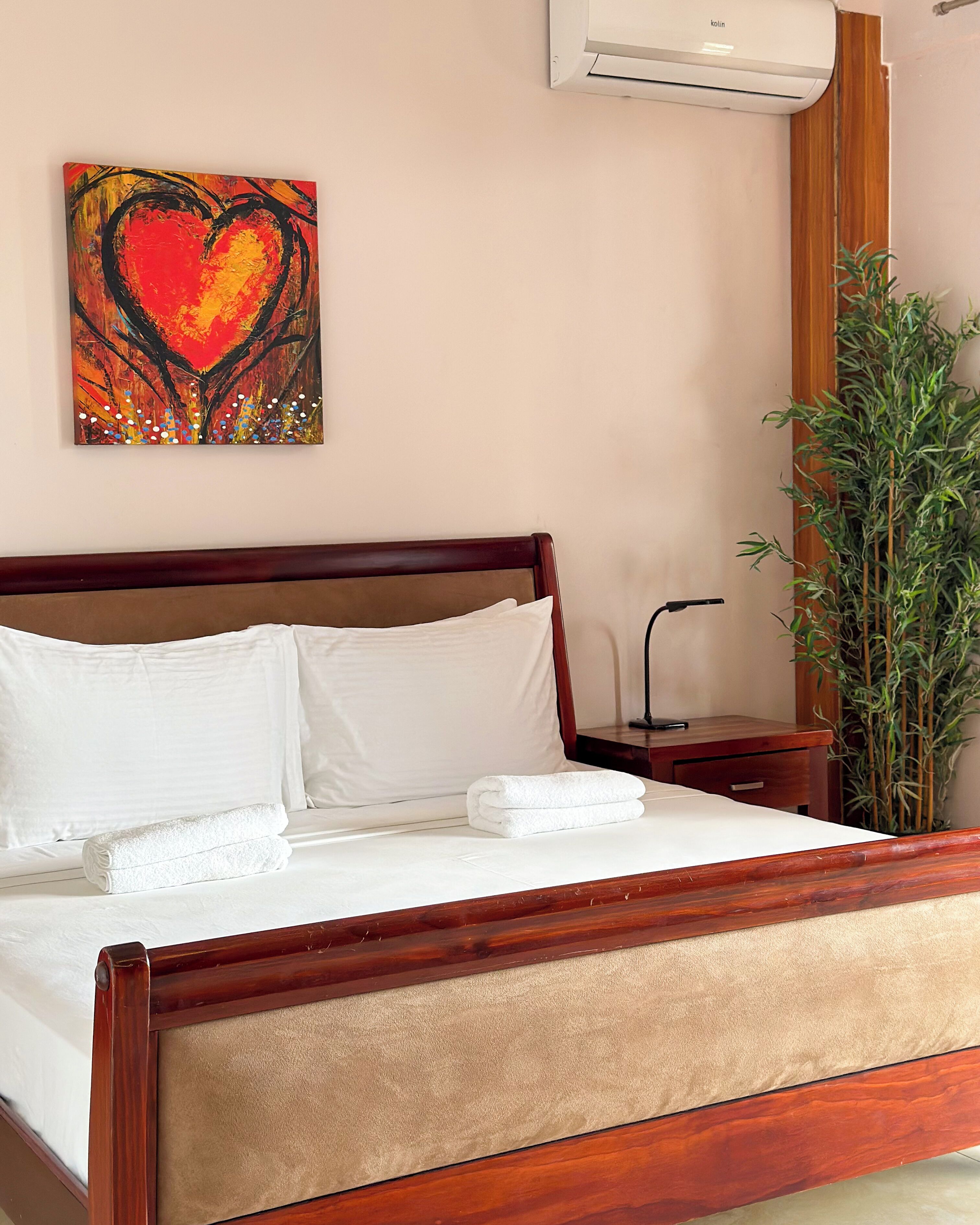 A neatly made bed with white linens and two towels, a wooden headboard, a wall-mounted air conditioner, and a tall green plant beside the bed.