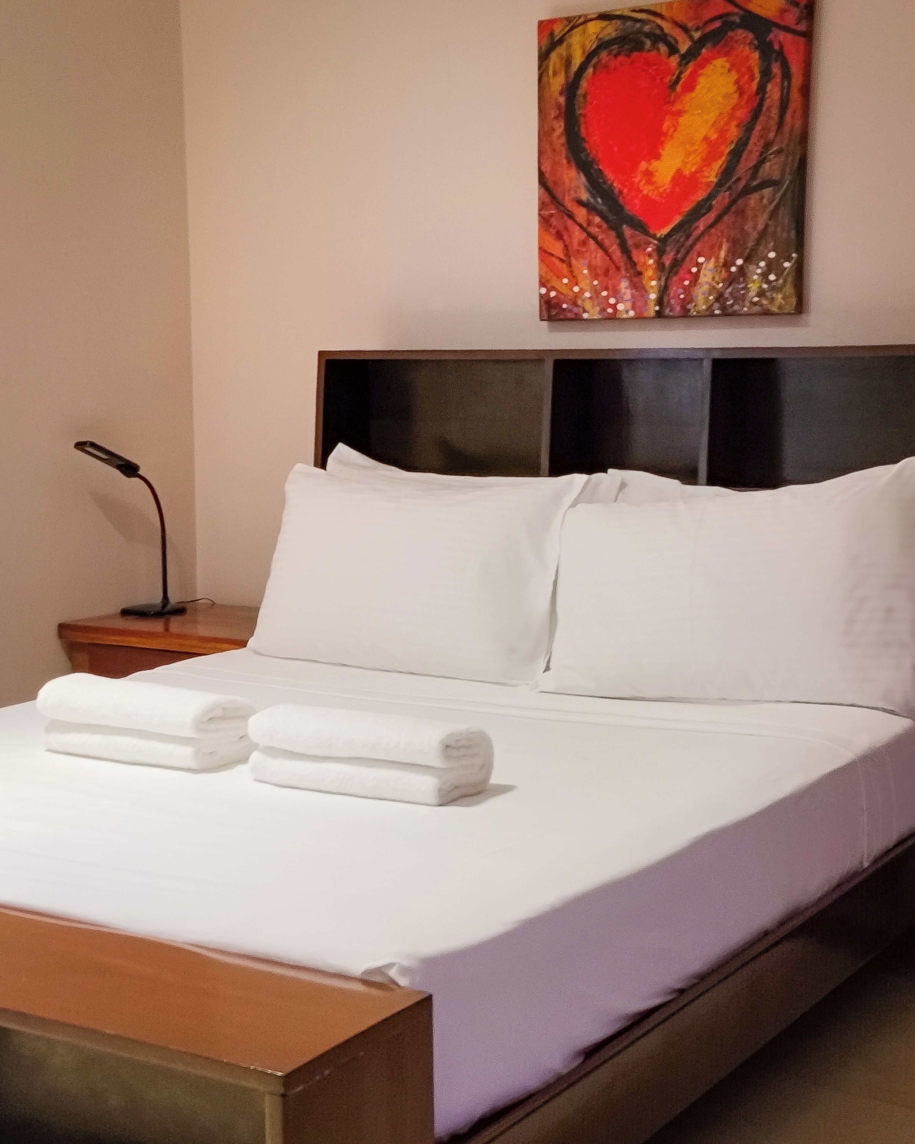 Una cama bien hecha con sábanas blancas y dos toallas enrolladas, una mesita de noche con una lámpara, y un cuadro en la pared con forma de corazón.