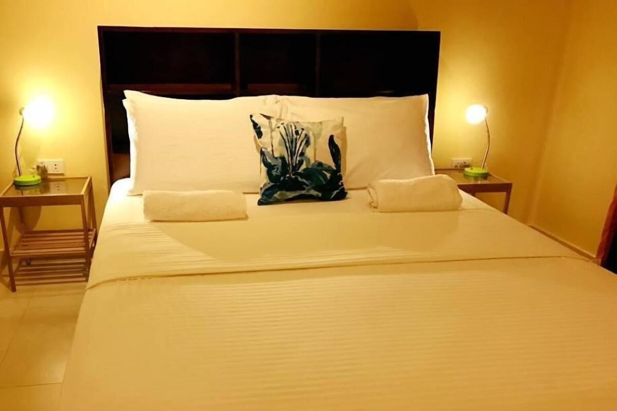 Una cama bien tendida con sábanas blancas, un cojín decorativo y dos lámparas de noche sobre mesitas de madera.
