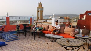 A rooftop terrace with a view of a tower, furnished with sofas, tables, and chairs. - Original Surf Morocco - Hostel (Aourir)