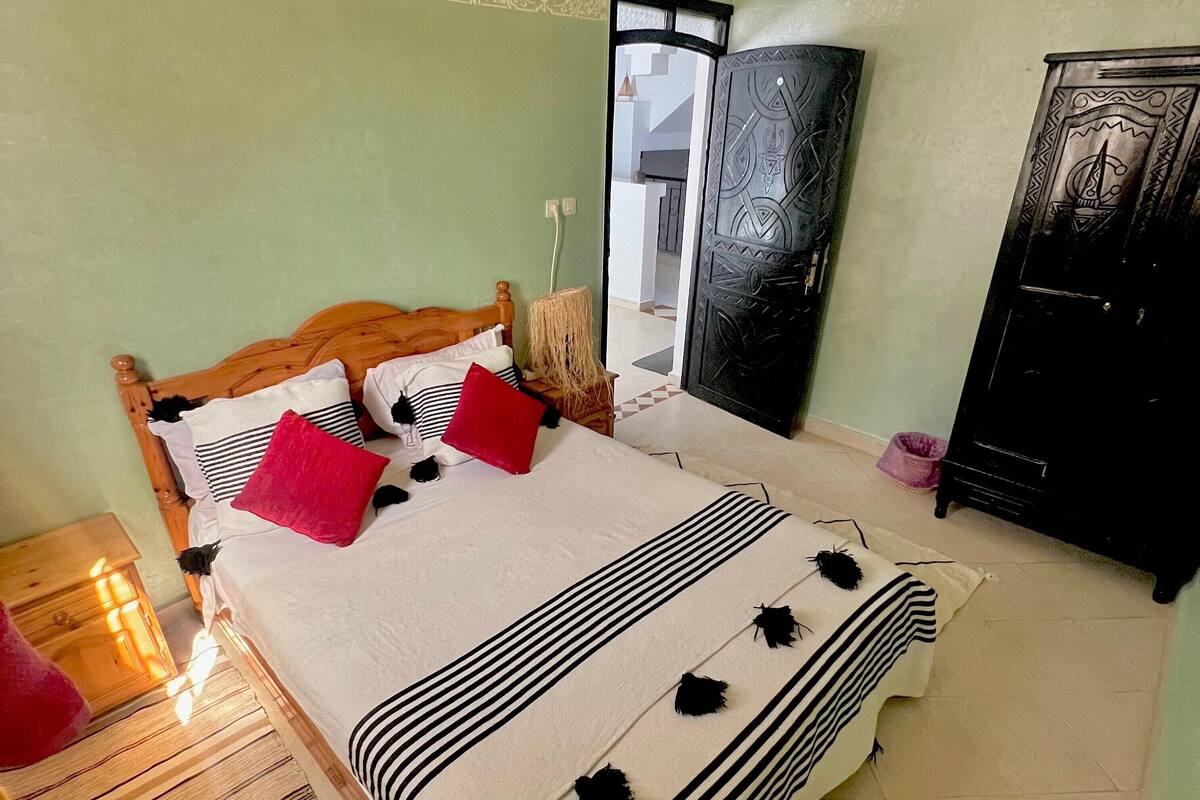 Uma cama bem arrumada com roupa de cama listrada e cabeceira de madeira, uma mesa de cabeceira com abajur, e um guarda-roupa encostado na parede.