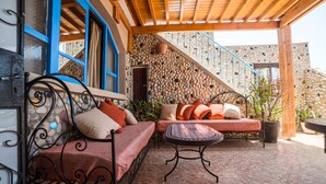 A cozy outdoor seating area with a wrought iron bench and a cushioned sofa, a stone wall with a mosaic design, and a wooden ceiling structure. - Original Surf Morocco - Hostel (Aourir)