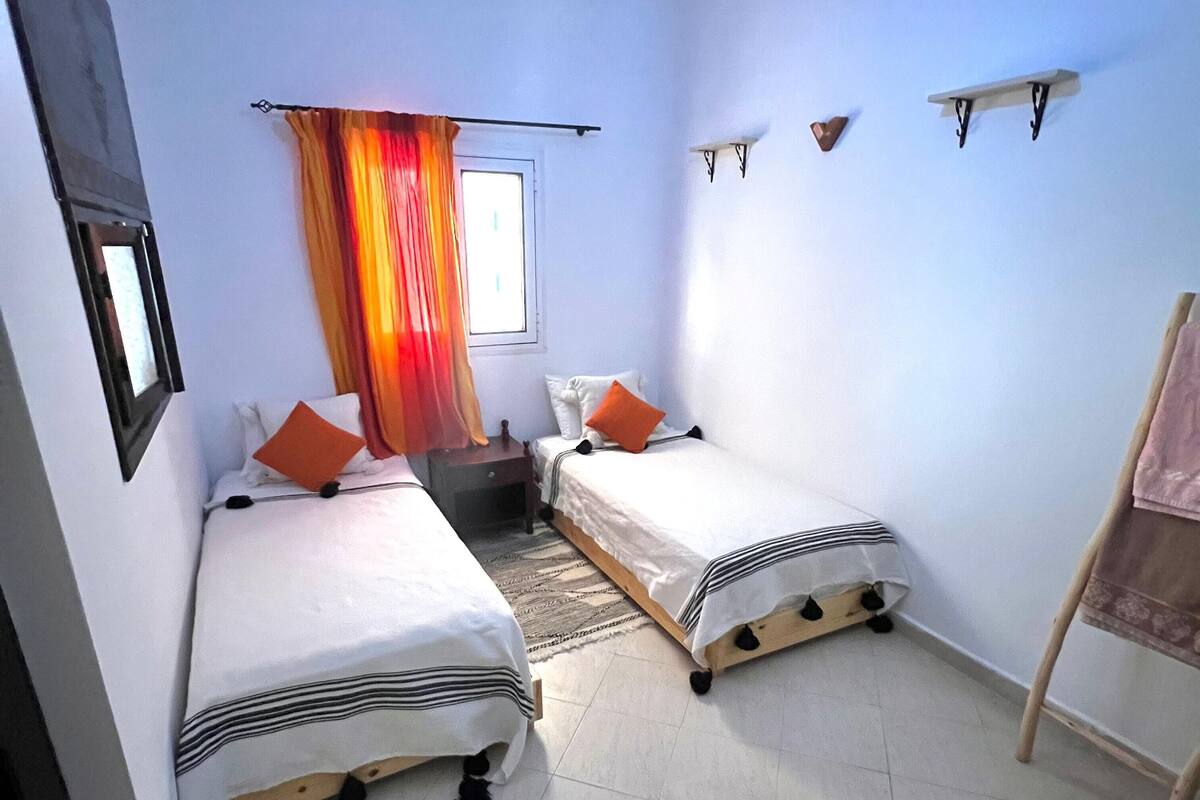 Quarto pequeno e bem iluminado, com duas camas de solteiro, uma mesa de cabeceira, janela com cortinas e um pequeno estante com objetos decorativos.