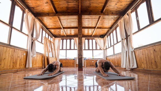 A room with wooden walls and ceiling, large windows with curtains, and two individuals on yoga mats. - Original Surf Morocco - Hostel (Aourir)