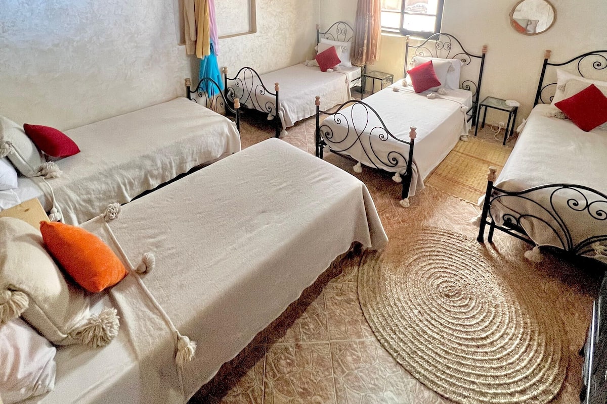 Quarto com quatro camas, tapete tecido, ventilador de teto e janela com cortinas.