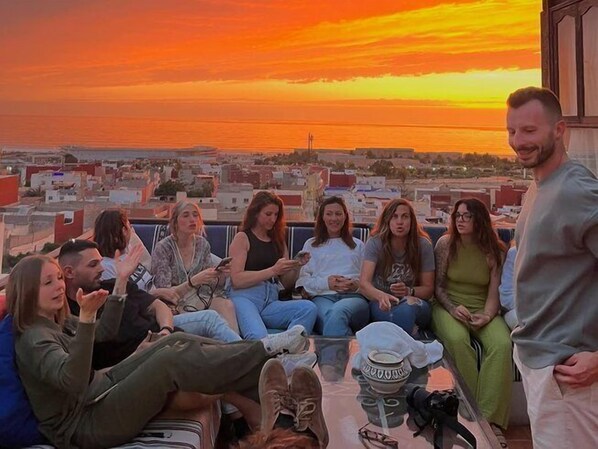 A group of people sitting on a balcony with a cityscape and sunset in the background. - Original Surf Morocco - Hostel (Aourir)