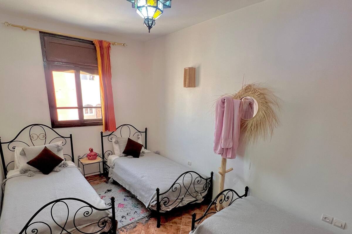 Quarto com duas camas, janela com cortinas, planta pendurada e piso de azulejos.