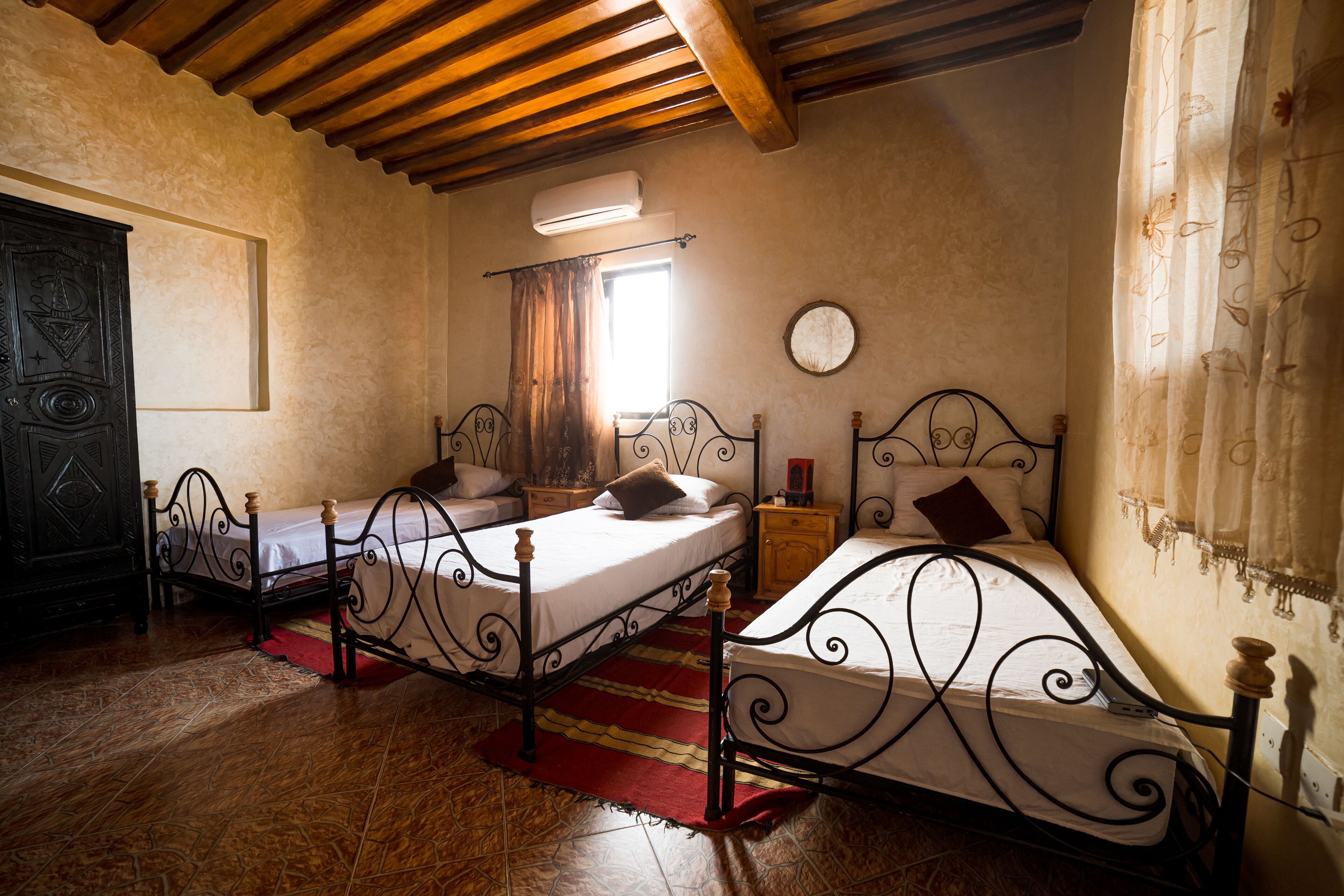 A room with two beds, a wooden ceiling, a window with curtains, a wall clock, and a small wooden nightstand.