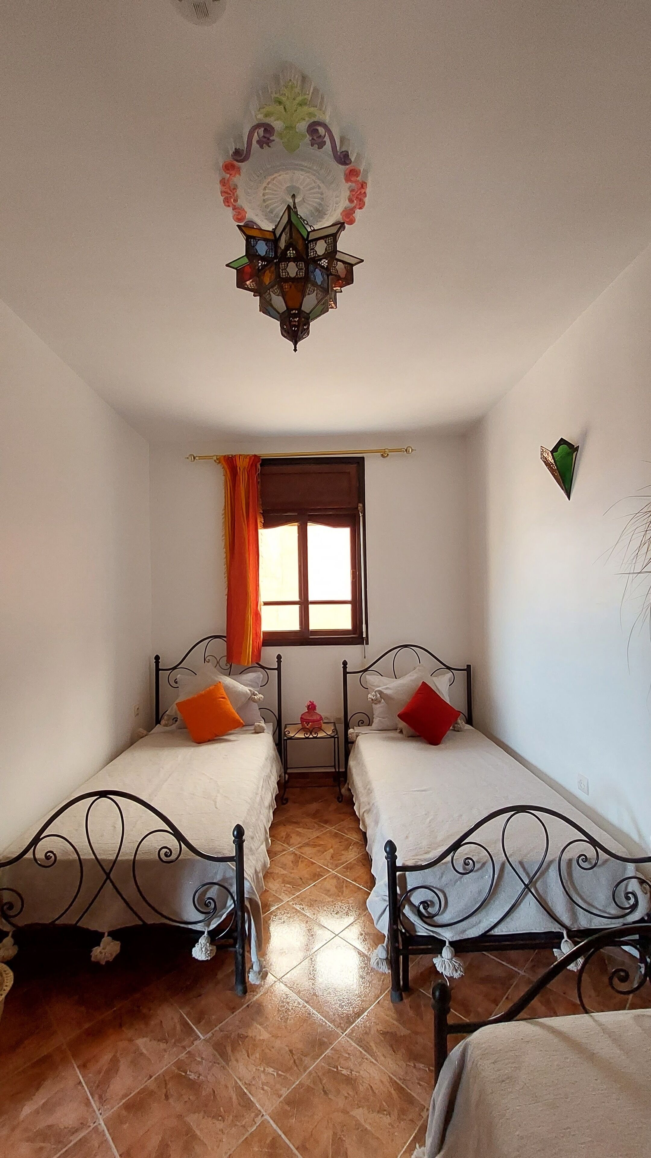 A room with two beds, a window with curtains, a decorative ceiling light, and a small table with a red object.