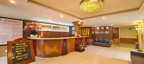 A hotel reception area with a wooden reception desk, a welcome sign, a chandelier, and a sofa. - HaiDuong Intourco Resort (Vung Tau)