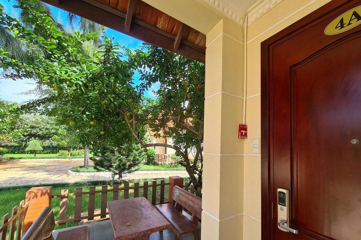 A wooden door with a number plate, a bench, and a table on a porch with a view of a tree and a building in the background.