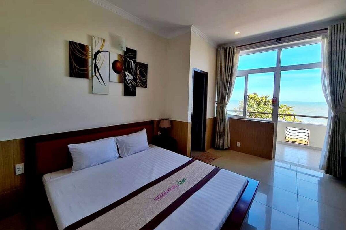 A hotel room with a large bed, a view of the ocean, and artwork on the wall.