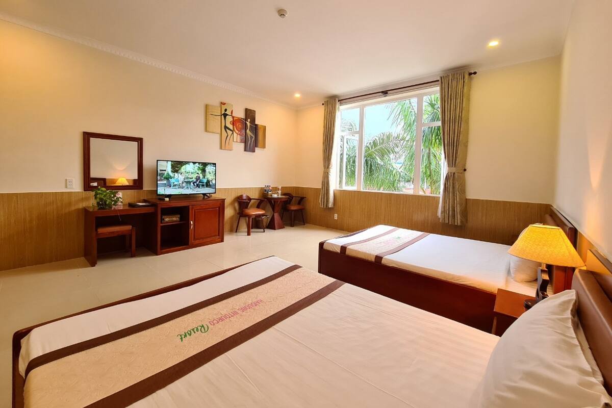 A hotel room with two beds, a desk with a TV, a window with curtains, and artwork on the wall.