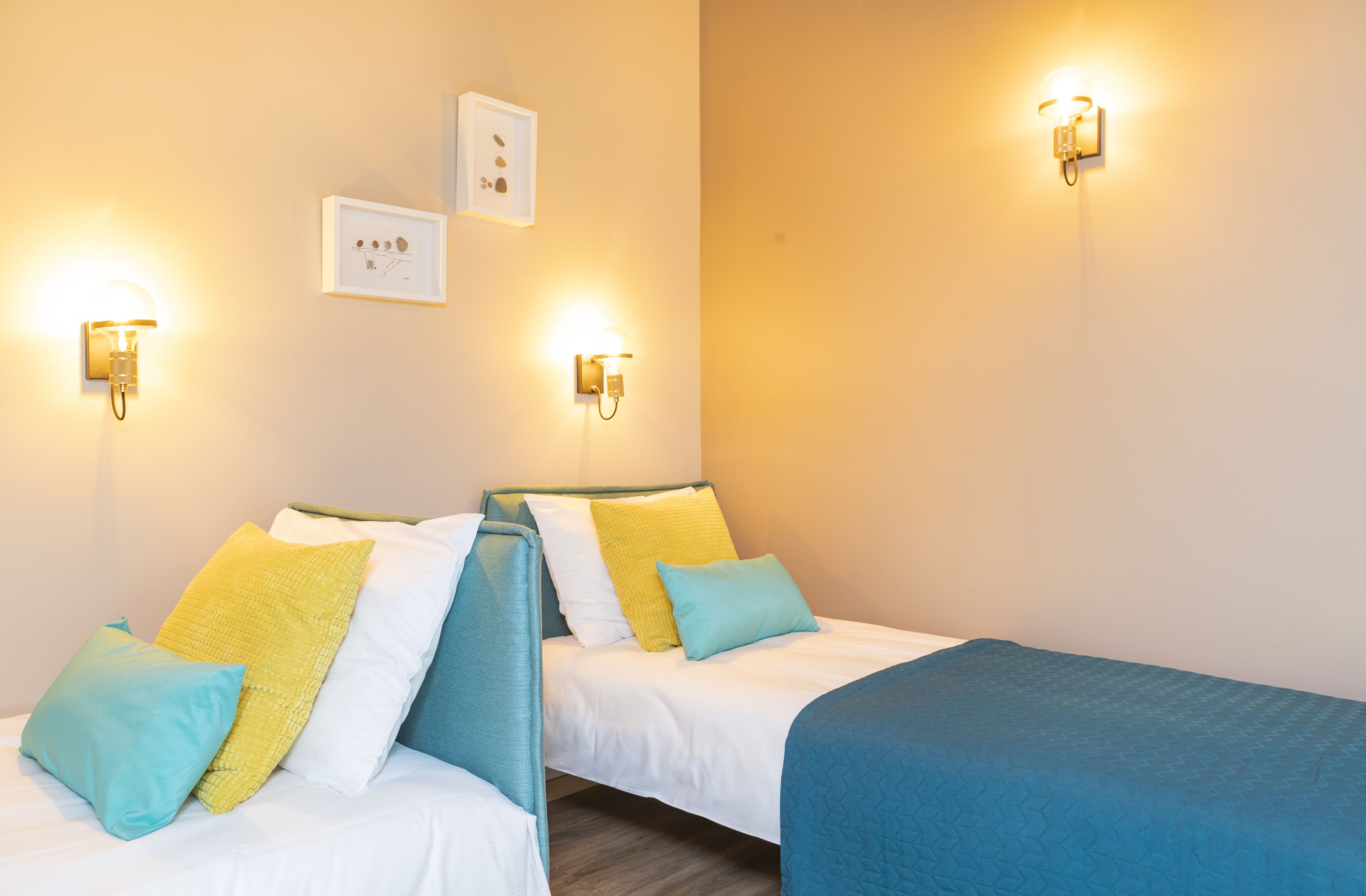 A room with two beds, each with a headboard, and two wall-mounted light fixtures. There are framed pictures on the wall.