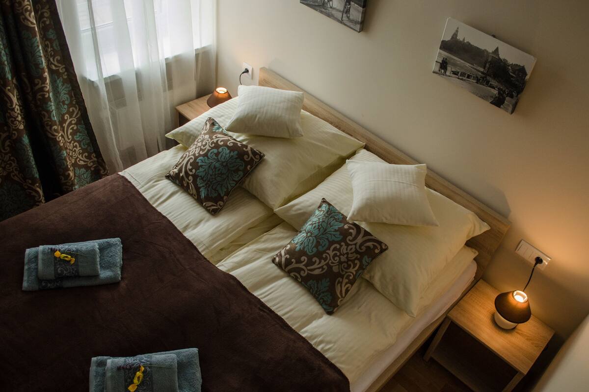 A neatly made bed with multiple pillows and a blanket, a bedside lamp, and a wall-mounted picture.