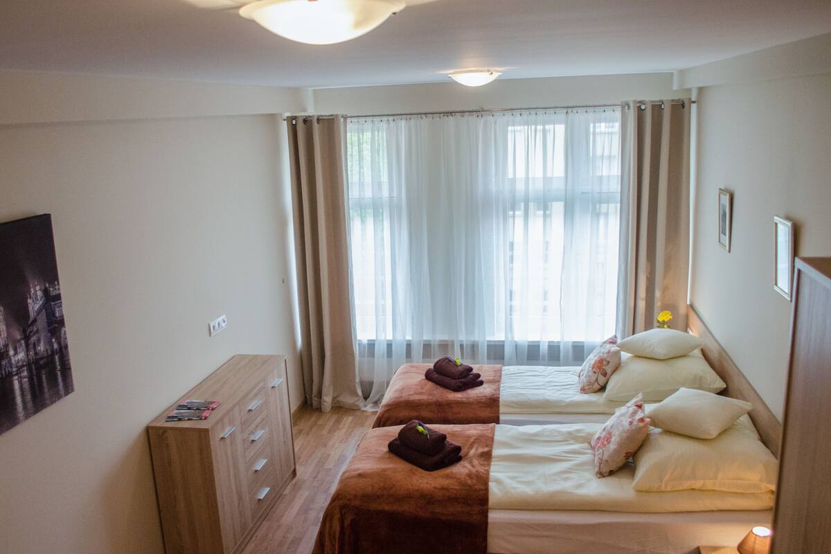 A hotel room with two beds, a wooden dresser, a nightstand, and a window with sheer curtains.
