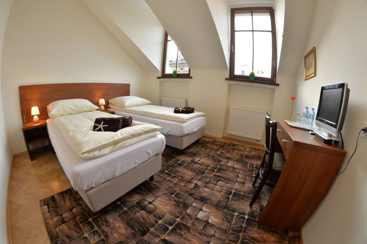 A bedroom with two beds, a desk with a computer, and a window with a view of buildings.