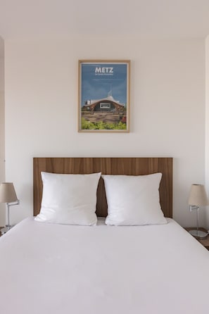 Quarto de hotel com cabeceira de madeira, duas almofadas brancas, um quadro emoldurado de um edifício na parede e duas luminárias de cabeceira.