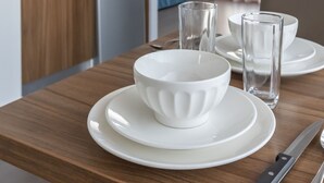 A modern kitchen with wooden cabinets, a microwave, and a coffee maker. There is a dining table with white plates, bowls, and glasses, and a knife on the table. - Odalys City Campus Apartment Hotel - Metz Centre (Metz)