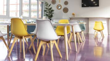 A modern dining area with a long table and chairs, large windows, and a flat-screen TV on the wall.