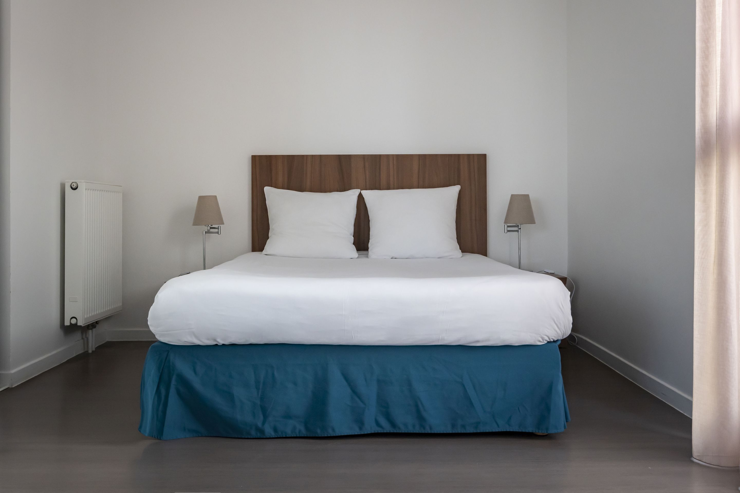 A neatly made bed with white linens and a blue skirt, a wooden headboard, two bedside lamps, and a white radiator.