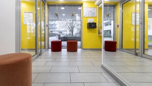 A reception area with a yellow wall, glass doors, and a tiled floor. There are three red ottomans and a brown chair. - Odalys City Campus Apartment Hotel - Metz Centre (Metz)