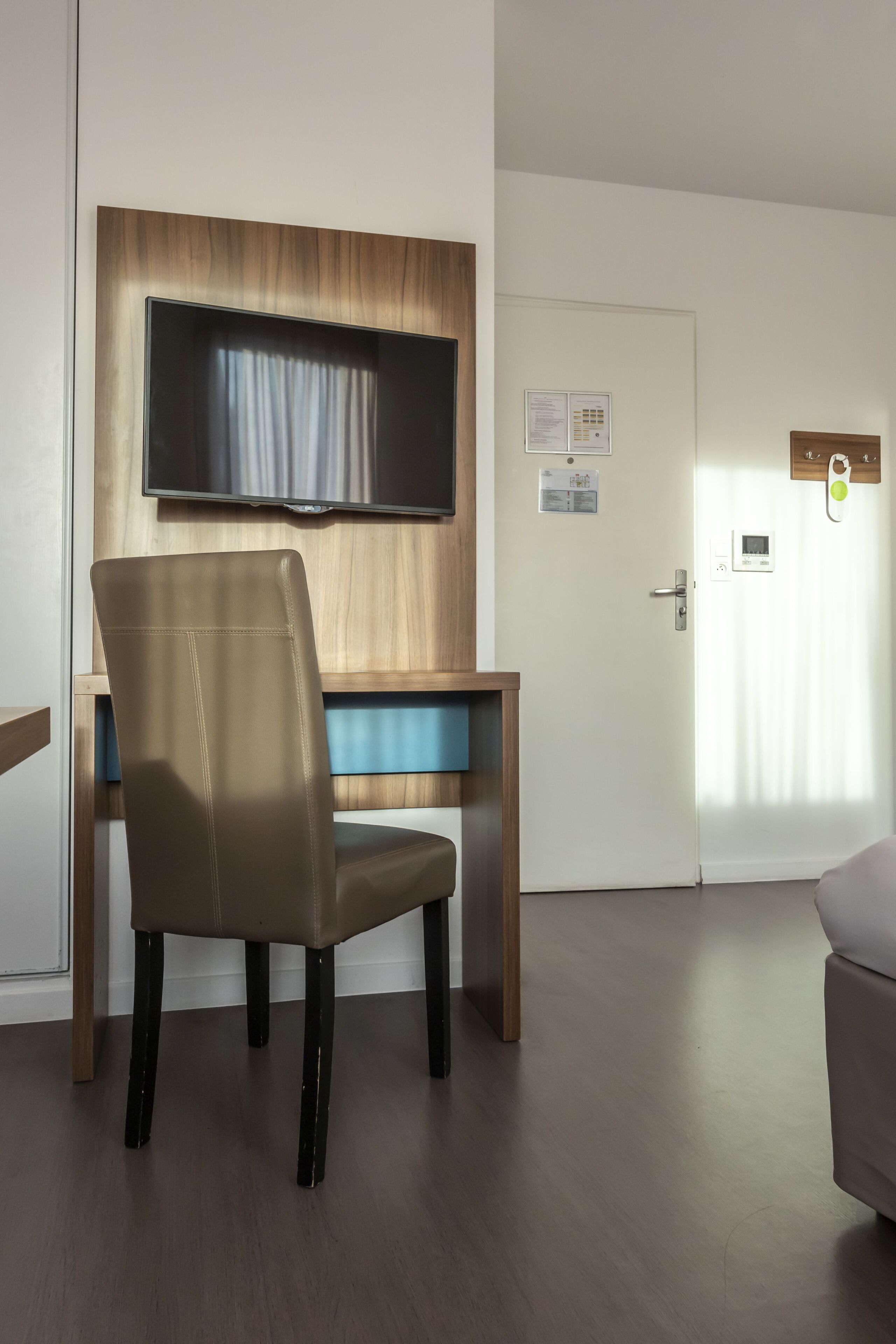 Een moderne hotelkamer met een houten bureau, een leren fauteuil, een flatscreen-tv aan de muur, een deur met een digitaal slot en een klein bedieningspaneel.