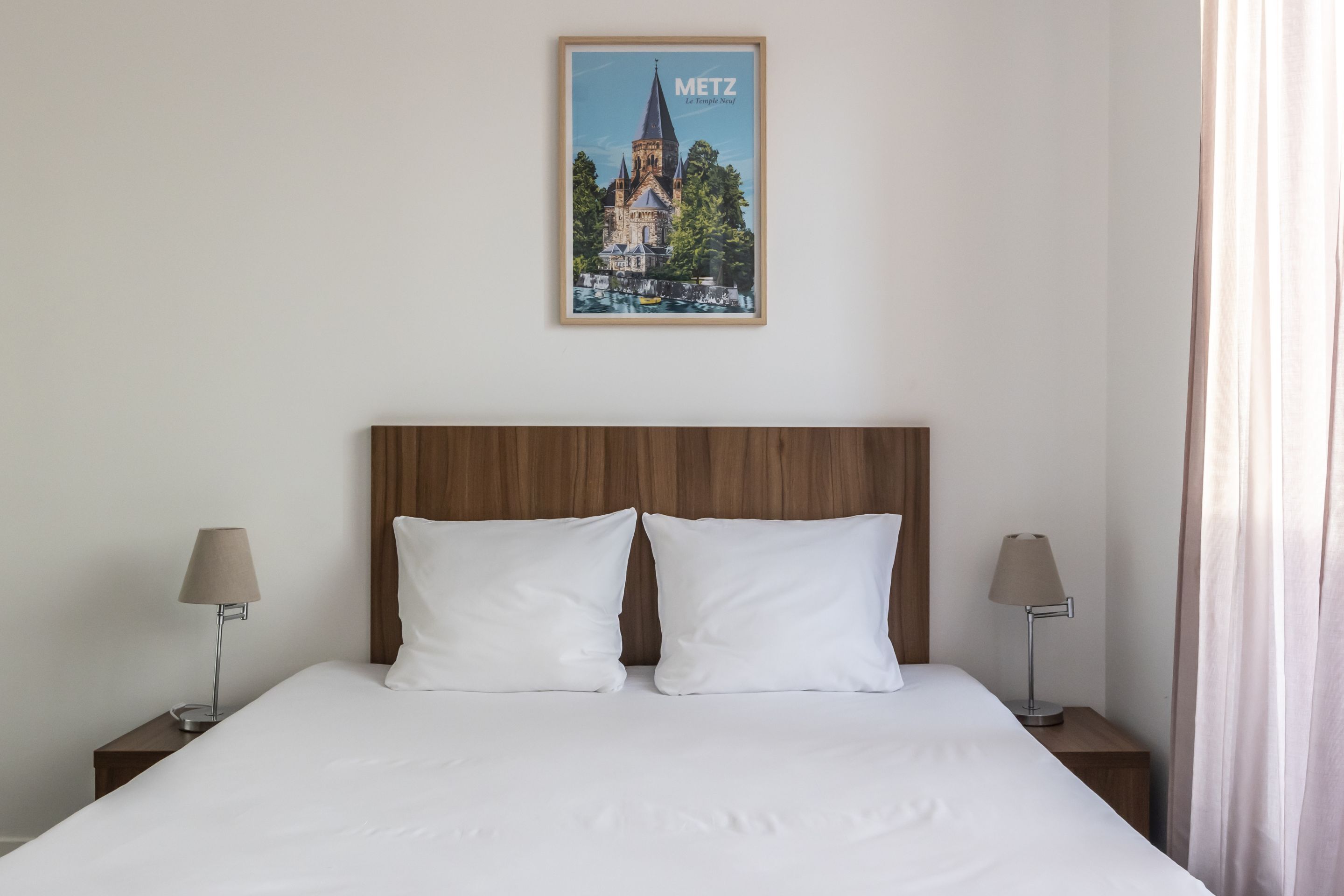 A neatly made bed with white linens, two bedside lamps, and a framed picture of a church on the wall.
