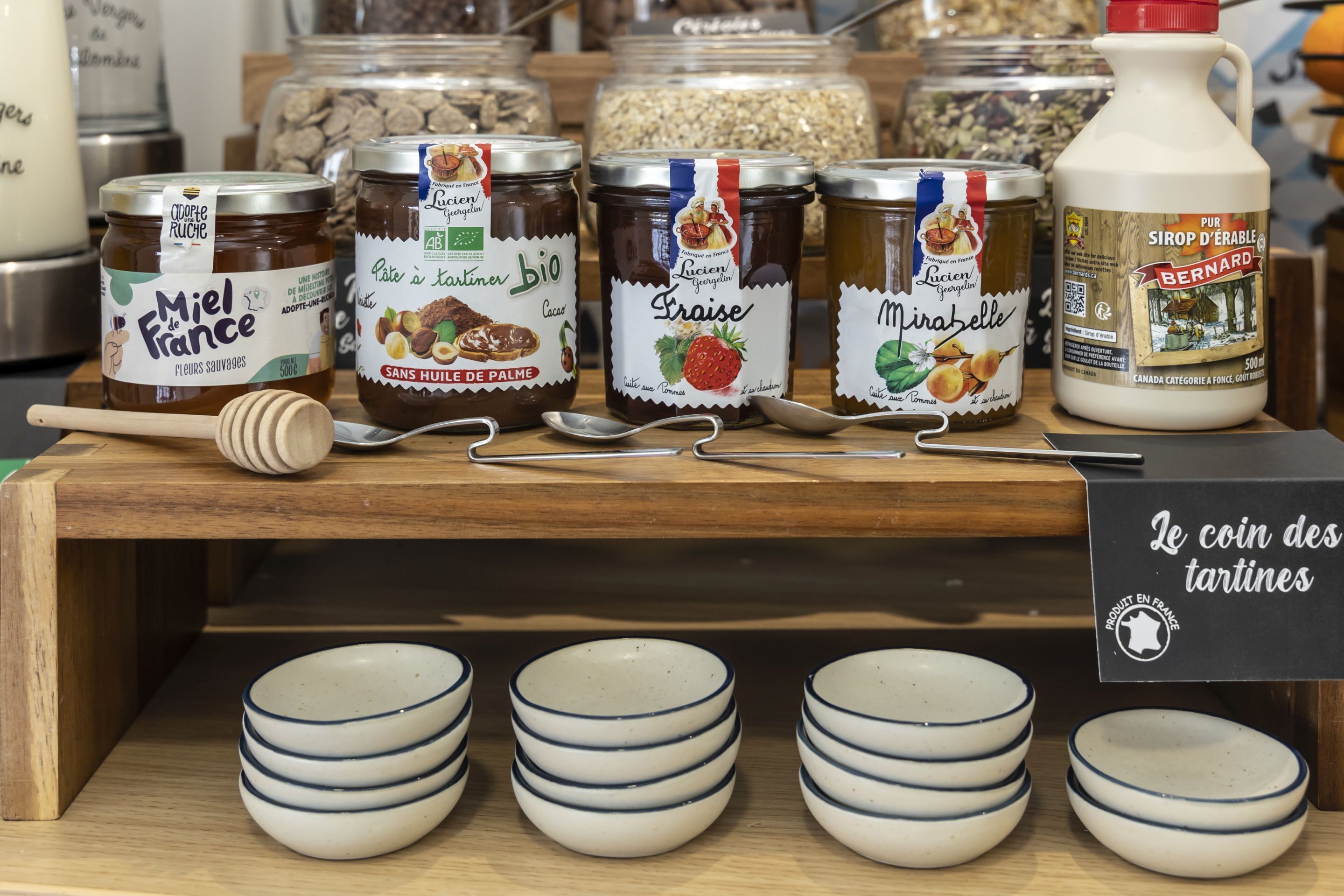 A display of various food products including jars of preserves and a bottle of milk, with small bowls and a sign indicating a coin-operated tartine machine.