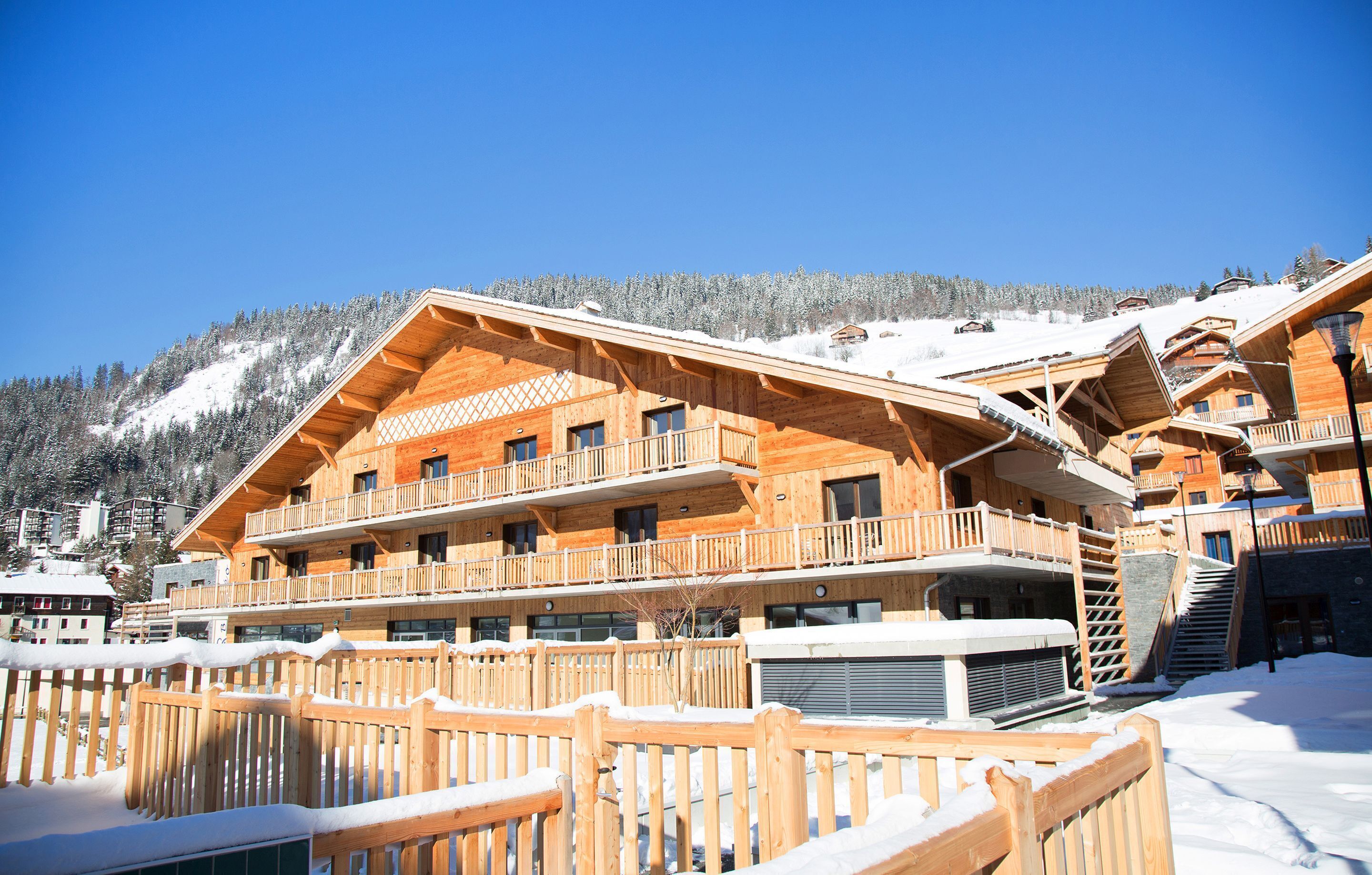 A wooden chalet with multiple balconies, a wooden fence, and a snowy mountain backdrop.