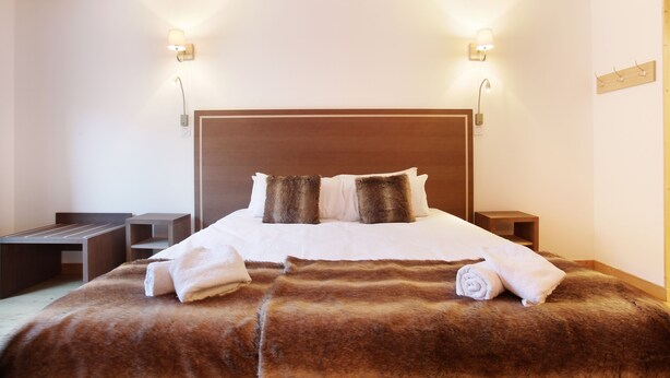 Ein ordentlich bezogenes Bett mit einer braunen Decke, zwei Kissen, einem Holz-Kopfteil und zwei Nachttischen mit Lampen.