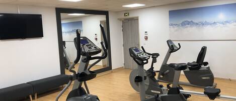A gym with three stationary bikes, a large mirror, a flat-screen TV, and a painting of mountains.