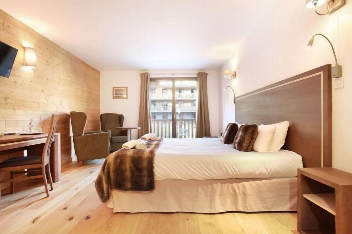 Une chambre d’hôtel avec un plancher en bois, un lit avec une literie blanche, un bureau avec une chaise, une petite table et un téléviseur fixé au mur.