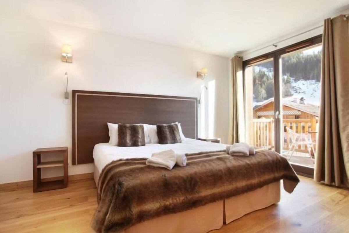 Une chambre avec un grand lit, une table de chevet, un plaid en fausse fourrure et une vue sur une terrasse en bois à l’extérieur.