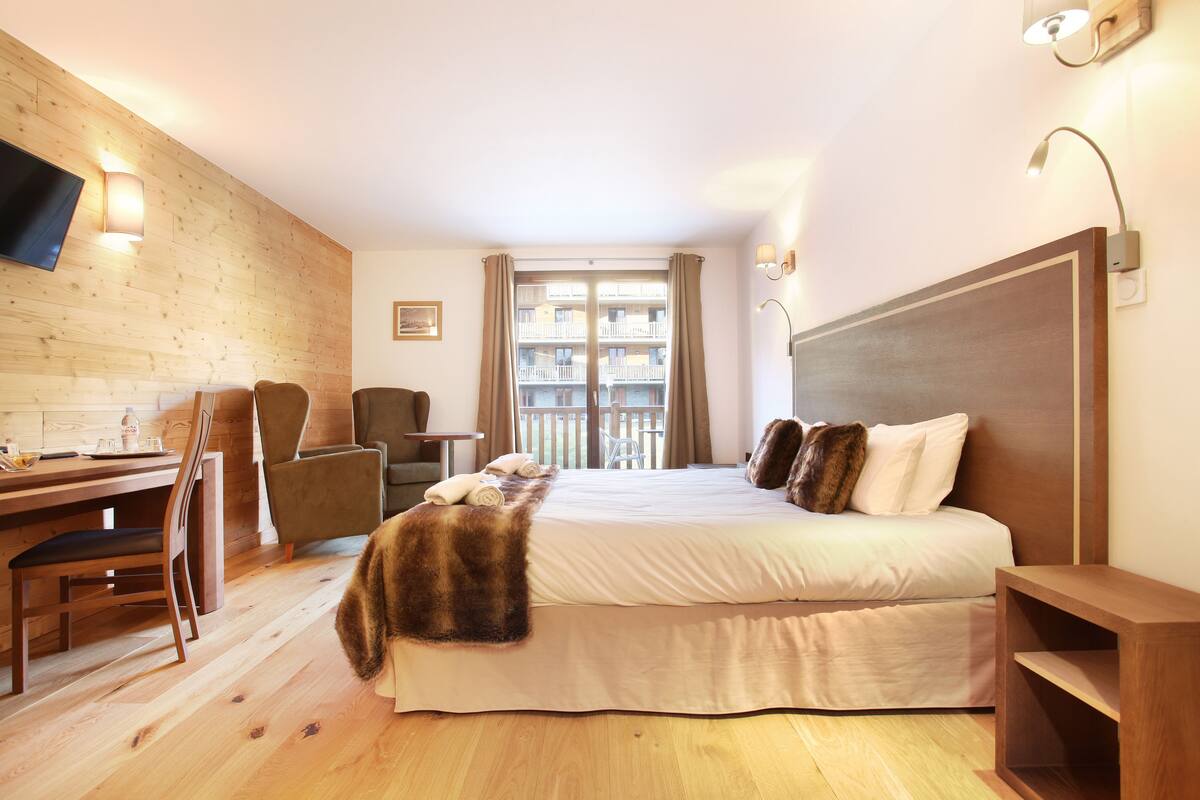Une chambre d’hôtel avec un plancher en bois, un lit avec une tête de lit, un bureau avec une chaise, un téléviseur fixé au mur et une fenêtre avec des rideaux.